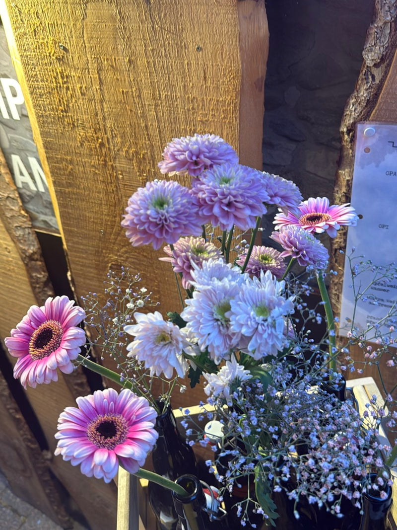 Diversi fiori di gerbera rosa si trovano insieme ad altri fiori in bottiglie davanti a una parete di legno.