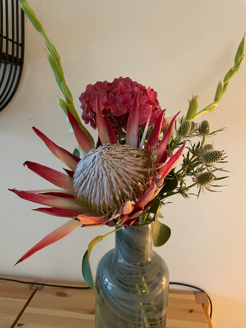 È visibile un grande fiore di protea reale (Protea cynaroides), che fa parte di un bouquet colorato in un vaso di vetro. Le caratteristiche brattee appuntite...